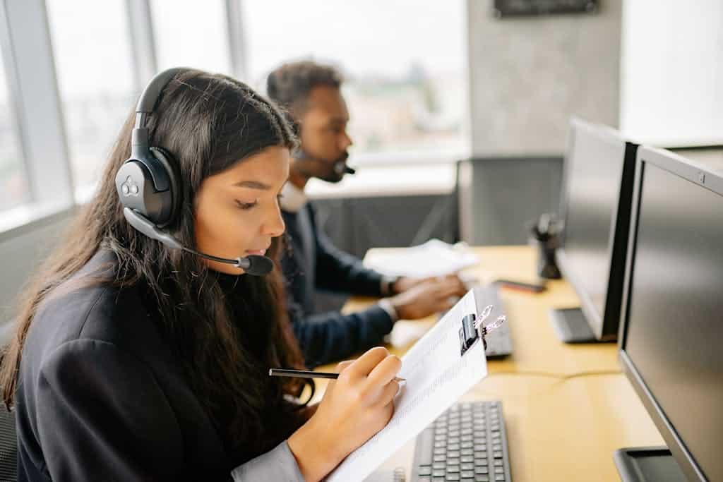 Two call center agents wearing headsets working at desks with computers in a modern office.