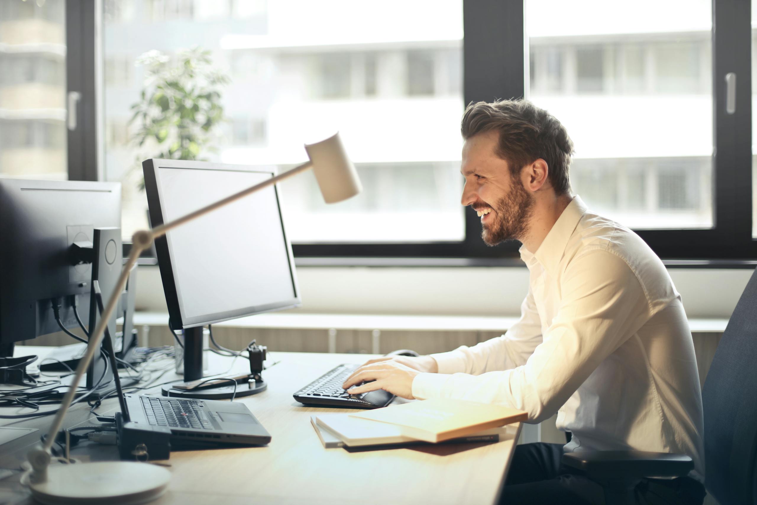 a man smiling while working at an office desk with a computer and natural daylight streaming in through large windows. 840996 scaled, Executive Search & Talent Acquisition