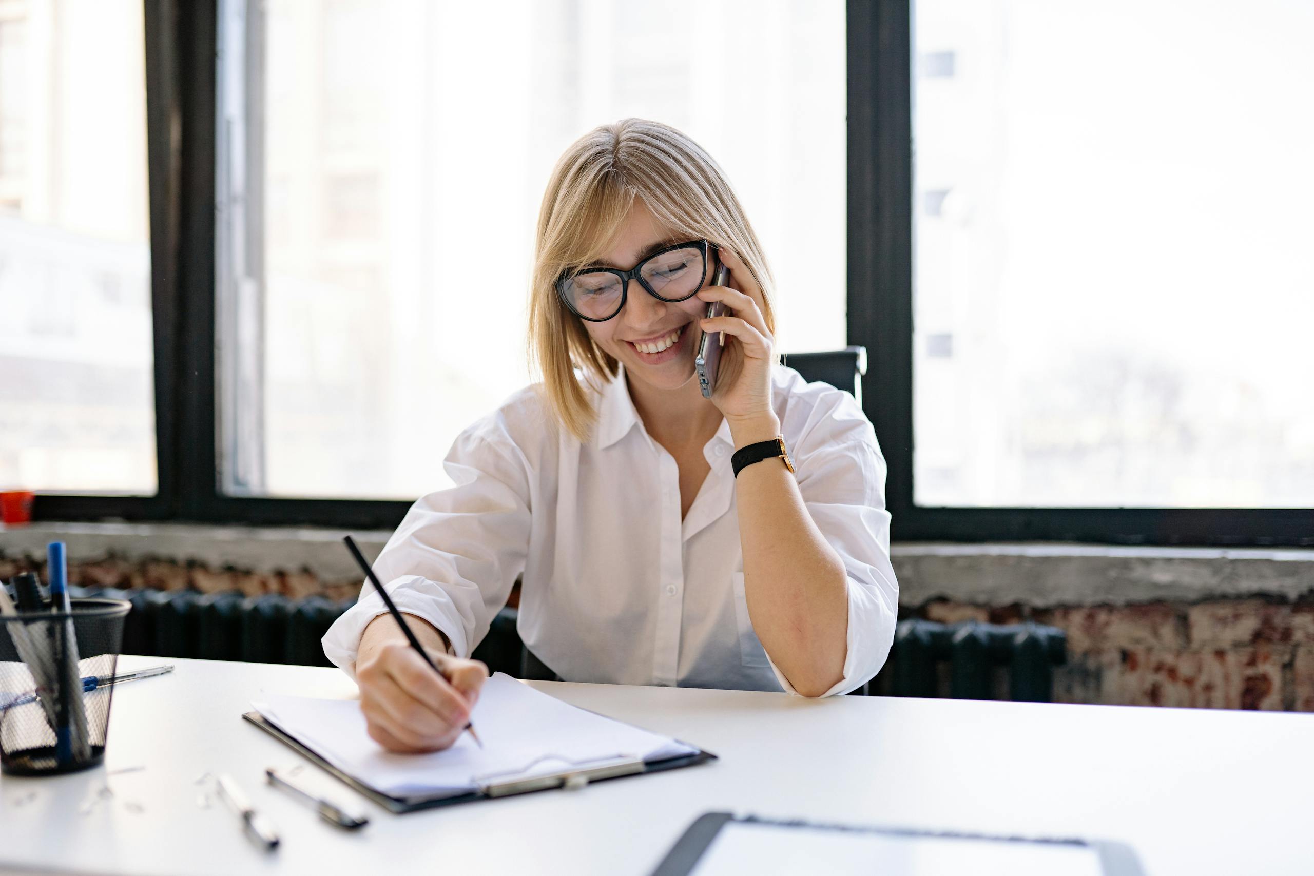 smiling woman talking on the phone while writing notes at a desk in a modern office. 7793187 scaled, TAG: Employment and HR Solutions